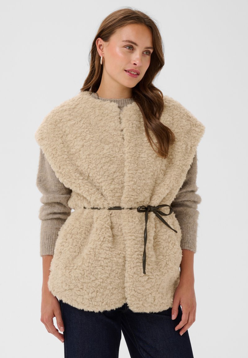 Γυναίκα φοράει μπεζ ψεύτικο γιλέκο με shearling, με μαύρη ζώνη-κορδόνι πάνω από γκρι πουλόβερ και σκούρο τζιν, μπροστά σε απλό φόντο.