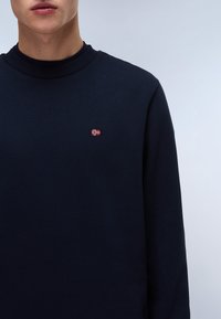 Marineblauwe sweatshirt met geribbelde halslijn, voorzien van een klein rood-wit vlaglogo op de borst. Zachte textuur, klassieke pasvorm, lange mouwen.