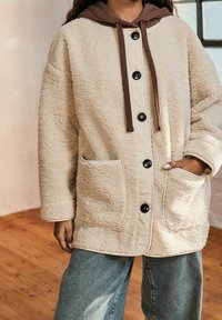 Giacca in shearling beige con cappuccio marrone, bottoni frontali e due tasche laterali. Texture morbida e design casual con una vestibilità comoda.