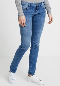 Hubené džíny ve světle modrém denimovém provedení se zesvětleným vzorem. Mají pět kapes, zipový patent a úzký střih. Kombinované s bílými teniskami.