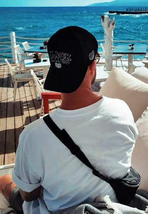 Black cap with "BLACK SKIES" embroidered, worn backward. White T-shirt, black sling bag, tattoo visible on left arm, wooden deck, ocean in background.