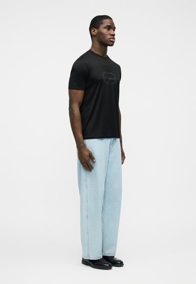 Άνδρας που στέκεται πλάγια φορώντας μαύρο μπλουζάκι με λογότυπο, ανοιχτό μπλε τζιν με φαρδιά πόδια και μαύρα επίσημα παπούτσια μπροστά σε λευκό φόντο.