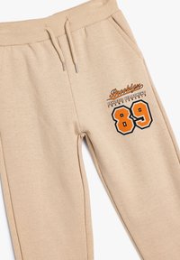 Pantalones de chándal beige hechos de tela suave, con una cintura ajustable con cordón, bolsillos frontales y un gráfico bordado de "Brooklyn 89".