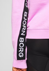 Sudadera de color rosa claro con panel de acento negro que presenta "BJÖRN BORG" en texto blanco a lo largo de la manga. Textura suave, diseño casual.