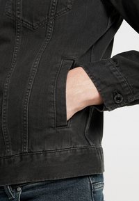 Giacca in denim nera con taglio classico, dotata di tasca sul petto e tasche laterali per le mani. Chiusure con bottoni sui polsini ben visibili.