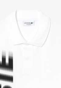Valkoinen polo-paita, jossa on teksturoitu kangas, kaksi nappia kauluksessa ja musta graafinen kuvio vasemmalla puolella. Lacoste-logo valkoisena.