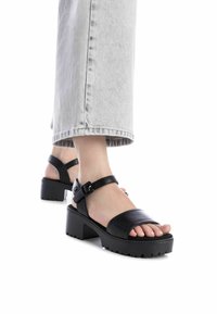 Sandalias de cuero negro con tacón grueso, correa ajustable en el tobillo y punta abierta. La suela texturizada presenta surcos profundos para un mejor agarre.
