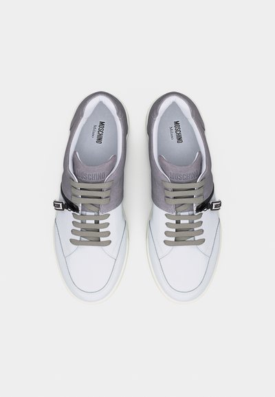 Ζευγάρι λευκά και γκρι αθλητικά παπούτσια Moschino Milano με μπεζ κορδόνια και μικρή μαύρη αγκράφα στο πλάι κάθε παπουτσιού.