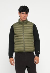 Olíva zöld pufi mellény magas gallérral, elülső cipzárral és Lacoste logóval. Fekete hosszú ujjú ing fölött viselve, bordás mandzsettákkal.
