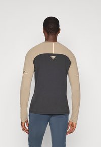 Hosszú ujjú póló, fekete testtel és bézs ujjal, hátul logóval és fényvisszaverő díszítéssel a középen. Sima anyagból készült.