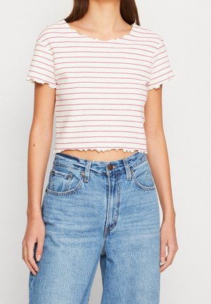 Vrouw die een wit cropped shirt met roze horizontale strepen en blauwe high-waisted jeans draagt, staand met armen ontspannen langs haar zij.