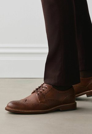 Braune Leder-Brogueschuhe getragen mit dunkelbrauner Hose, zeigen detaillierte Nähte und perforierte Muster auf einem hellen Boden.