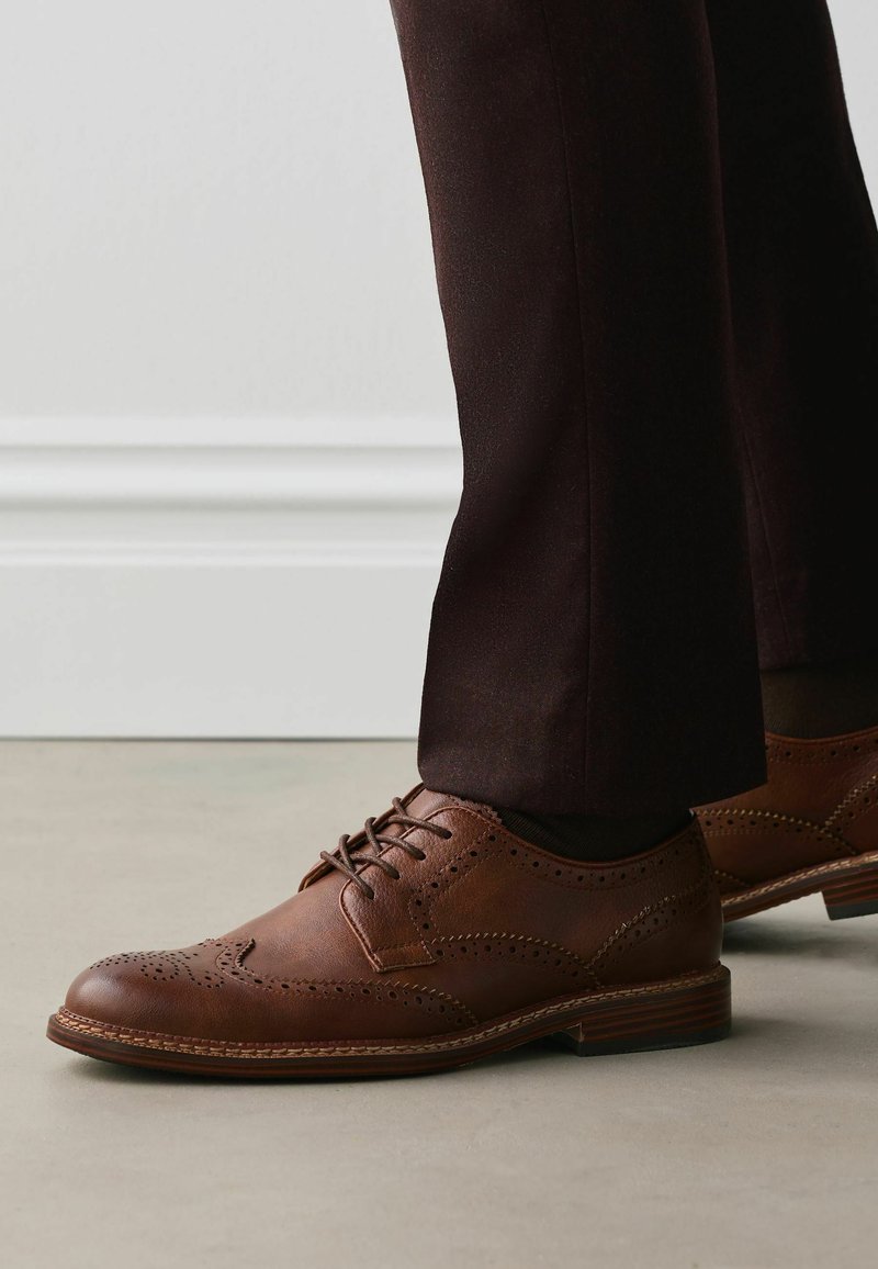 Chaussures brogues en cuir marron portées avec un pantalon marron foncé, montrant des coutures détaillées et des motifs perforés sur un sol clair.