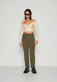 Top corto beige a coste con spalla scoperta e dettaglio intrecciato, abbinato a pantaloni cargo verde oliva con tasche laterali, indossati con sneaker nere.