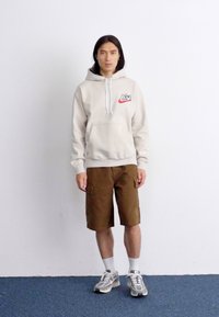 En person står op ad en ensfarvet hvid væg, iført en beige hættetrøje og brune shorts, med sportssko og en neutral ansigtsudtryk.