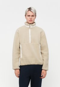 Beige fleecegenser med kvart glidelås, høy krage og hvite detaljer. Har sidelommer; myk tekstur, avslappet passform.
