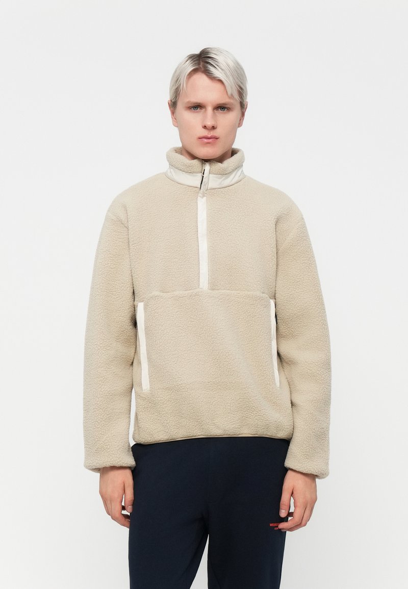 Beige fleecegenser med kvart glidelås, høy krage og hvite detaljer. Har sidelommer; myk tekstur, avslappet passform.