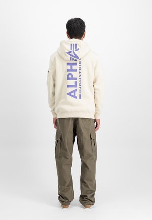 Homme portant un sweat à capuche crème avec un grand logo vertical "Alpha Industries" dans le dos, associé à un pantalon cargo olive et des chaussures décontractées, debout de dos.
