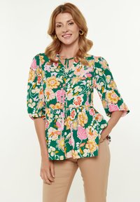 Top a peplo floreale verde con motivi floreali rosa, arancioni e gialli. Presenta un colletto a chiave e maniche puff a tre quarti.