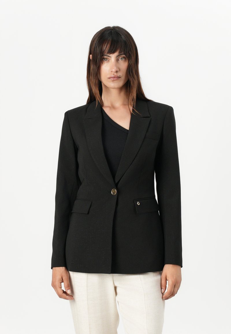 Blazer nero con una vestibilità sartoriale, due tasche frontali, chiusura con un bottone singolo e un tessuto leggermente strutturato. Abbinato a un top nero e pantaloni chiari.