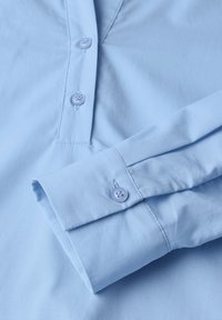 Camicia azzurra chiara con una pattina abbottonata dotata di quattro bottoni coordinati, maniche lunghe con polsini abbottonati e una texture liscia.