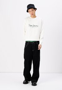 Sudadera blanca con logo verde de "Pepe Jeans", pantalones holgados negros, sombrero de cubo negro y zapatos gruesos negros. Patrón de tablero de ajedrez visible.