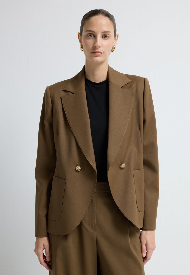 Ruskea kaksinkertainen bleiseri, joka on valmistettu sileästä kankaasta. Siinä on näyttävät olkatyynyt, kultasävyiset napit ja sivutaskut, ja se on yhdistetty mustaan yläosaan.