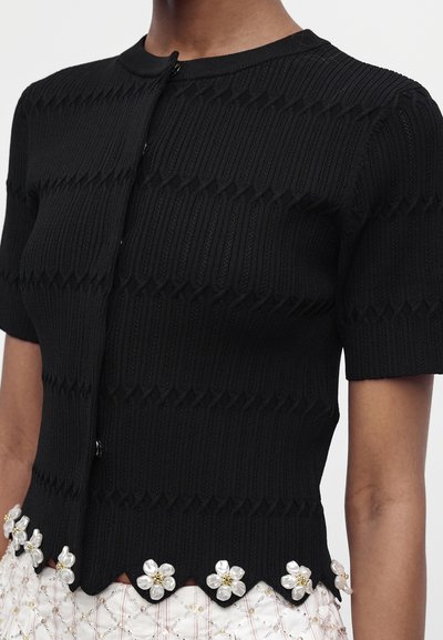 Haut en maille noir à manches courtes avec un ourlet festonné décoré d'embellissements floraux en perles et or, porté avec une jupe claire à motifs.