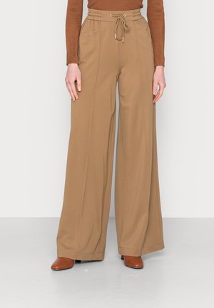 Vrouw draagt beige wijde broek met hoge taille en trekkoord, gecombineerd met bruine schoenen met puntige neus en een geribd topje.