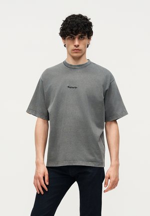 Νέος άνδρας με σγουρά μαλλιά που φοράει ξεθωριασμένο γκρι φαρδύ μπλουζάκι με το λογότυπο "Replay Inc." και σκούρα τζιν, στέκεται μπροστά σε απλό φόντο.