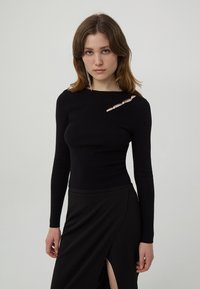 Czarna, prążkowana bluzka z długim rękawem z wycięciem i dekoracyjnymi akcentami. Dopasowany krój uzupełnia czarna spódnica z rozcięciem na udzie.