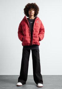 Chaqueta acolchada roja con capucha, con cierre de cremallera y detalle del logo. Se lleva sobre una camiseta de manga larga negra y se combina con pantalones de pierna ancha negros.