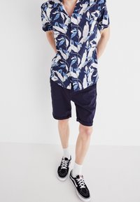 Košile s krátkými rukávy s modro-bílým vzorem listů, spárovaná s námořnickými šortkami. Model má na sobě černé tenisky s bílou podešví a ponožky.