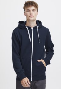 Tamno plavi dukserica na zip s mekim materijalom, s prednjim džepom, bijelim vezicama i rebrastim manžetama. Glatka tekstura, ležeran dizajn.