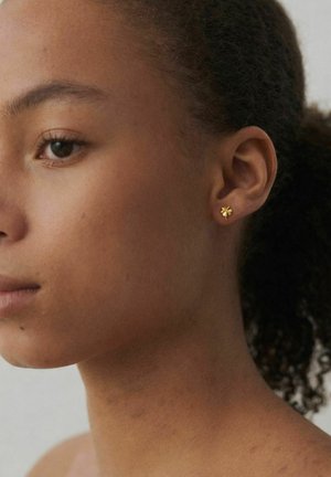 Mlada ženska z kodrastimi lasmi v nizkem čopku nosi majhne zlate uhane v obliki štiriperesne deteljice na levi ušesi na svetli podlagi.