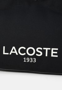 Černá plátěná taška se silným bílým nápisem "LACOSTE" a pod ním "1933", s texturovaným materiálem a prošívanými okraji.