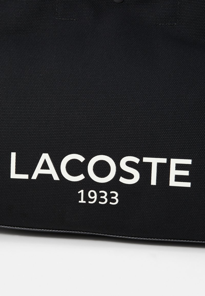 Borsa a tracolla nera con il logo "LACOSTE" in bianco in caratteri grandi e "1933" sottostante, realizzata in tessuto testurizzato e con bordi cuciti.