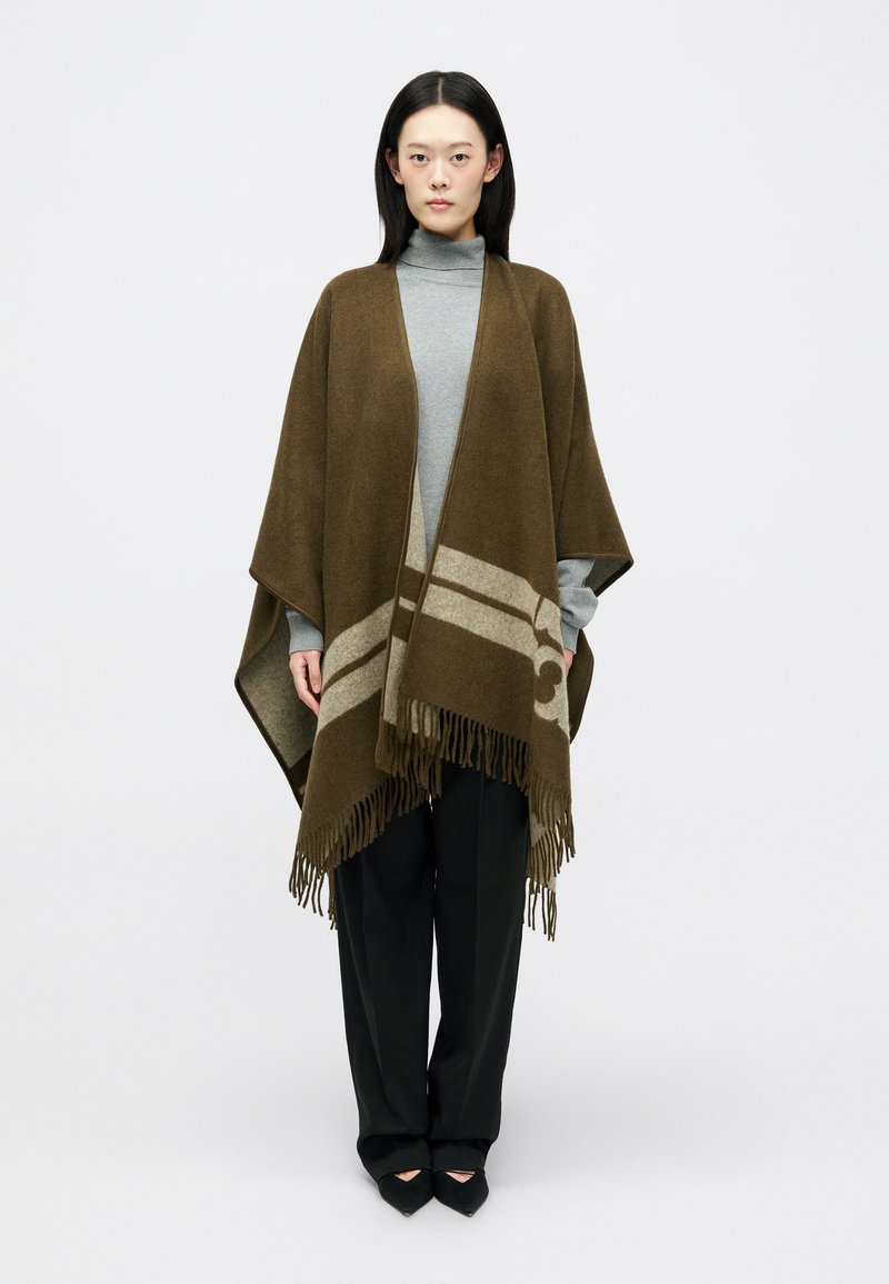 Poncho de lã castanho com uma faixa horizontal cinza e franjas. Usado sobre um pulôver cinza claro, combinado com calças e sapatos pretos.