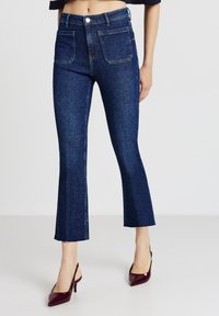 Jeans in denim a zampa d'elefante di colore blu scuro con vita alta, due tasche frontali e orlo grezzo. Abbinati a décolleté bordeaux con punta affusolata.