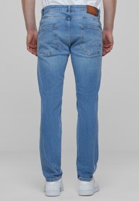 Jeans de mezclilla azul claro con corte recto, cinco bolsillos y un parche de cuero marrón en la cinturilla. Textura suave y color uniforme.