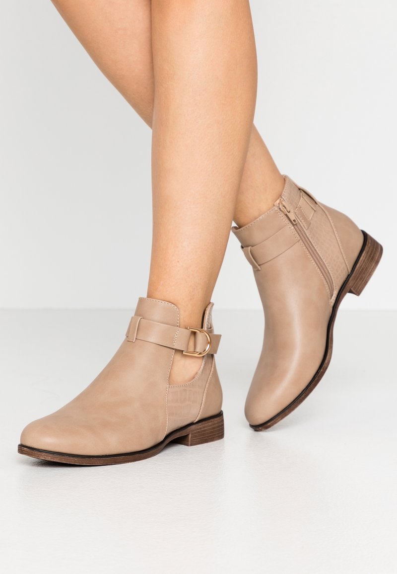 Jambes portant des bottines beige avec fermeture éclair latérale, sangle et boucle dorée, posées sur un sol blanc sur fond uni.