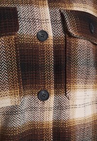 Bruin en beige geruite stof met twee zwarte knopen en een knoopzakflap op een gestructureerd shirt of jasje.