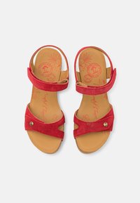 Sandales en suède rouge avec deux brides réglables, design ouvert au niveau des orteils, et une semelle intérieure en cuir marron ornée de motifs floraux. Détail en bouton doré inclus.