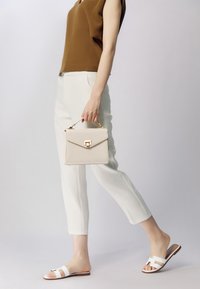 Jasno beżowa torebka z teksturowanym wykończeniem i złotymi akcentami, w zestawieniu z białymi, skrojonymi spodniami i białymi klapkami. Brązowy top dopełnia całość.