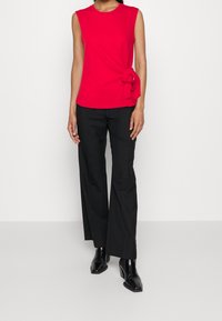 Top rojo sin mangas con cuello redondo, que presenta un detalle de nudo lateral. Combinado con pantalones de pierna ancha negros y botas de tacón negras.