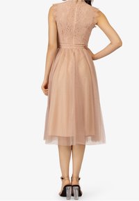 Femme portant une robe midi sans manches en dentelle et tulle rose poussiéreux avec une fermeture éclair au dos, assortie à des sandales à talons blocs transparentes.