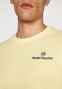 Sárga pamut póló kerek nyakkal, sötétlila logóval és "Sergio Tacchini" márkanevével a mellkason.