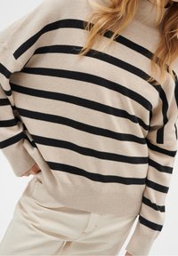 Pull beige avec des rayures horizontales noires, col rond et manches longues. Texture douce, coupe légèrement ample, avec poignets et ourlet côtelés.