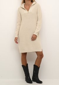 Femme portant une robe pull beige en tricot avec un col demi-fermeture éclair et des bottes de cowboy noires mi-mollet, debout contre un mur blanc.