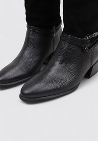 Botas de tobillo de cuero negro con un acabado texturizado, punta afilada, tacón bajo y recto, y una correa decorativa con acentos.